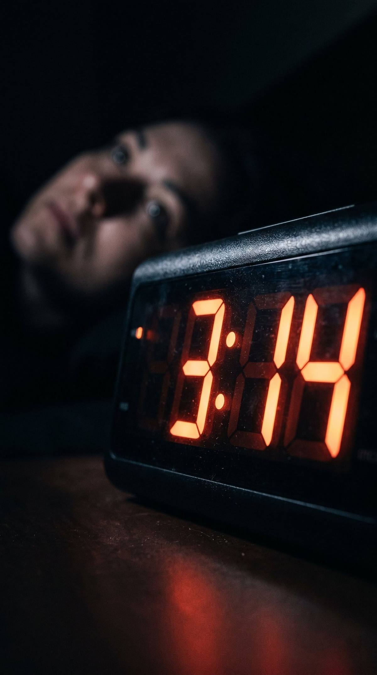 Digital alarm clock showing 3:14 AM, person visible in blurred background